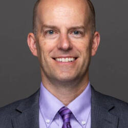 A man in a grey suit and purple shirt and tie smiles in to camera